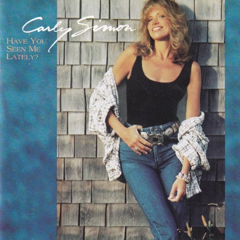 Carly Simon posiert lächelnd vor einer Holzverkleidung, trägt lässige Kleidung und wirkt entspannt.