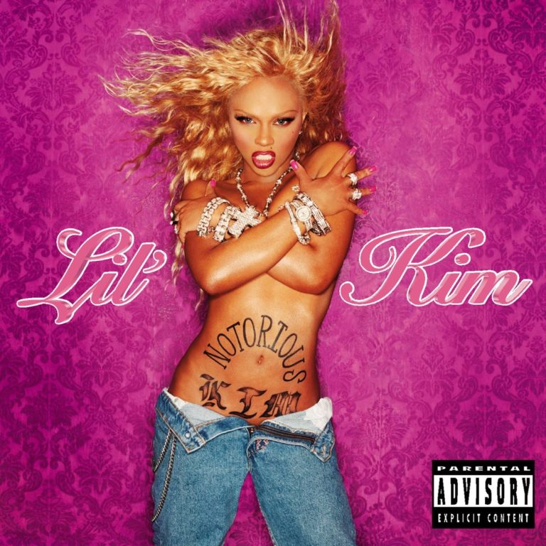 Lil Kim mit verschränkten Armen, freiem Oberkörper und Jeans vor pinkem Hintergrund.