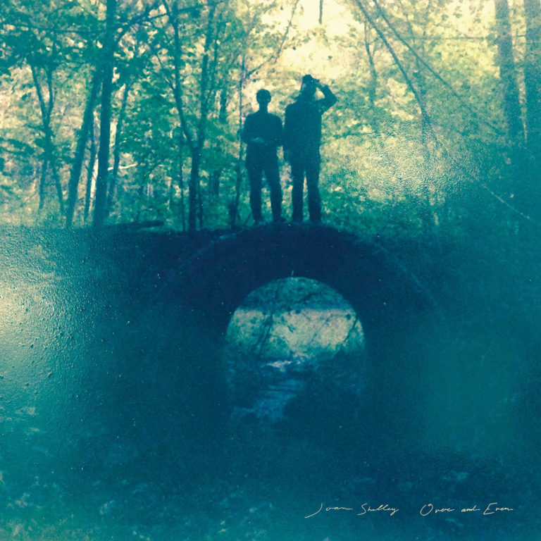 Zwei Personen stehen auf einer Brücke im Wald, umgeben von Bäumen und natürlichem Licht.