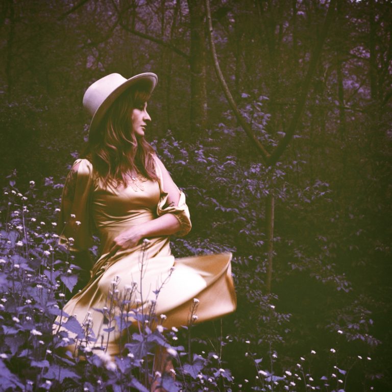 Frau in gelbem Kleid und Hut sitzt in einem Wald voller Blumen.