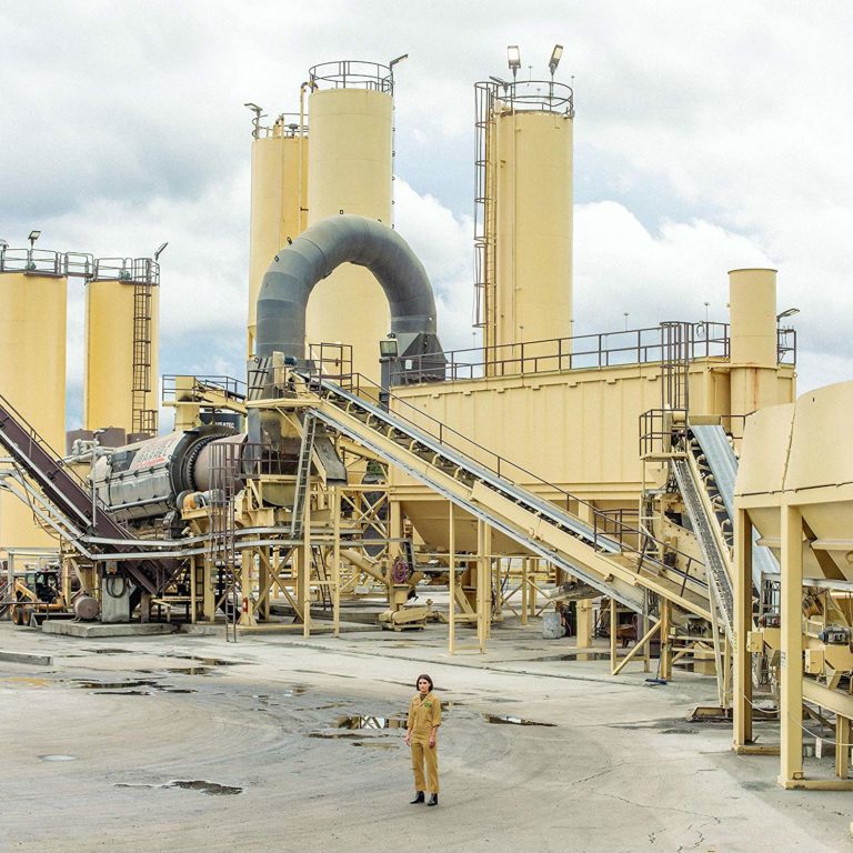 Person in gelbem Overall steht vor einer großen Industrieanlage mit Rohren und Silos.