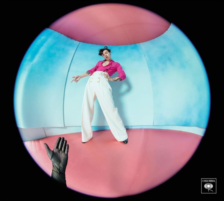 Person in pink shirt and white pants in a colorful, distorted room with a gloved hand in the foreground.