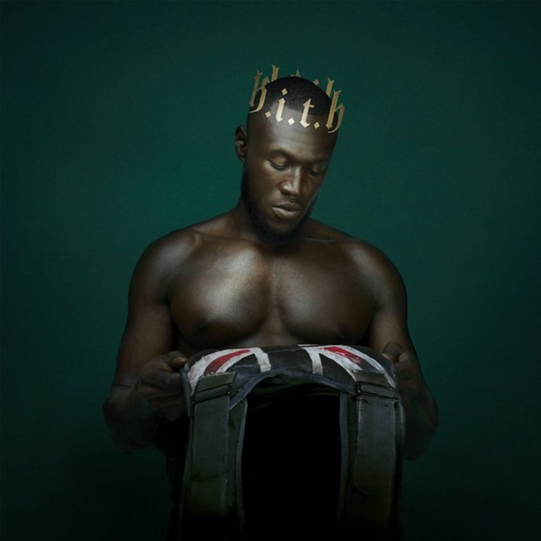 Stormzy steht oberkörperfrei mit einer Krone und einer Schutzweste in der Hand vor einem dunklen Hintergrund.
