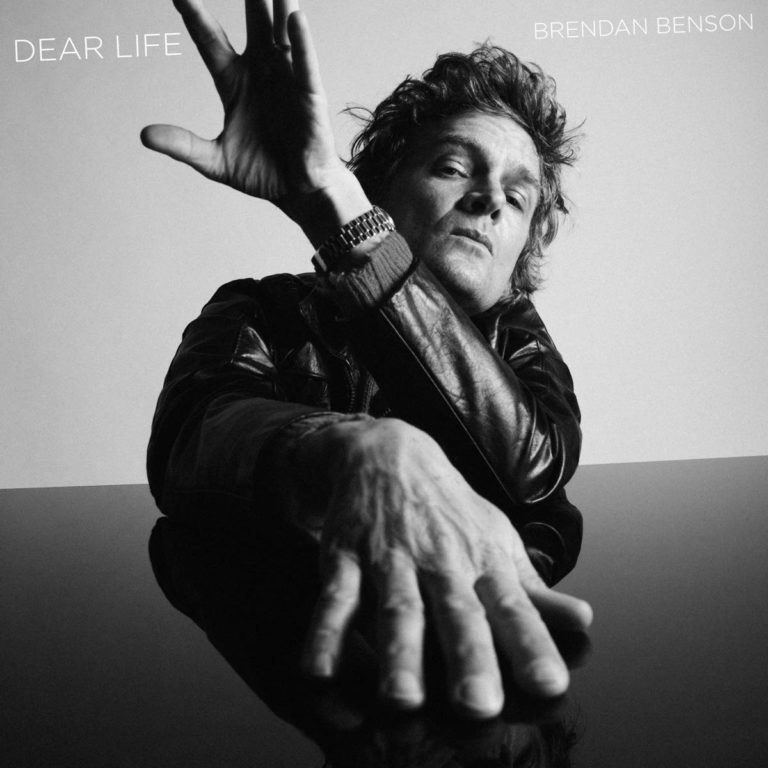 Schwarz-weiß Porträt von Brendan Benson mit ausgestrecktem Arm auf einem spiegelnden Tisch.
