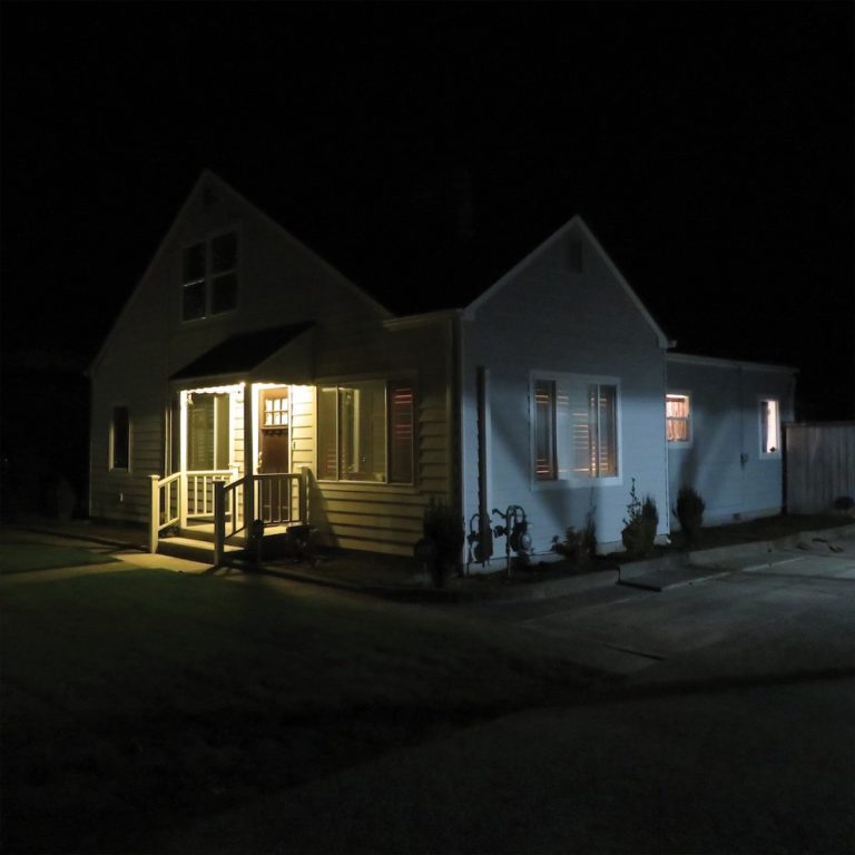 Ein beleuchtetes Haus bei Nacht mit Veranda und Fenstern, umgeben von Dunkelheit.