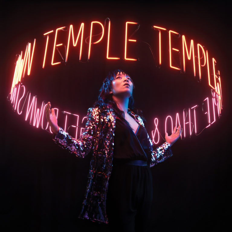 Person in glitzerndem Outfit vor neonfarbenem Schriftzug TEMPLE auf dunklem Hintergrund.