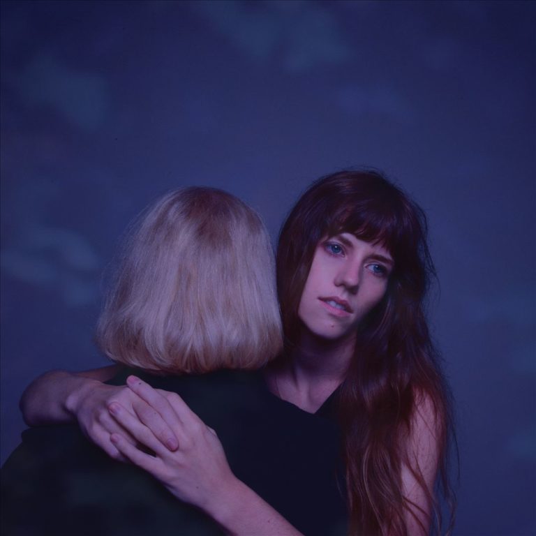 Frau mit langen Haaren umarmt Person mit blondem Haar vor blauem Hintergrund.
