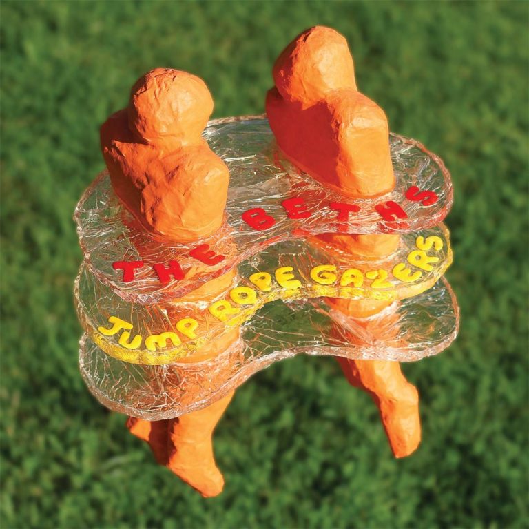 Zwei orange Figuren aus Pappmaché stehen auf grünem Gras, umgeben von transparenten Schichten mit Schriftzug.