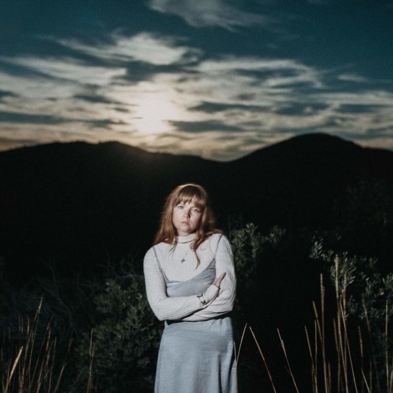 Frau in heller Kleidung steht vor einer Berglandschaft bei Sonnenuntergang.