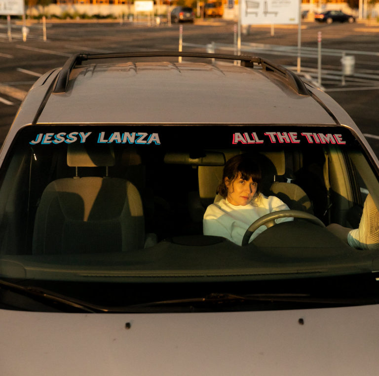 Jessy Lanza sitzt im Minivan, ein Fuß auf dem Armaturenbrett, bei Sonnenuntergang auf einem leeren Parkplatz.