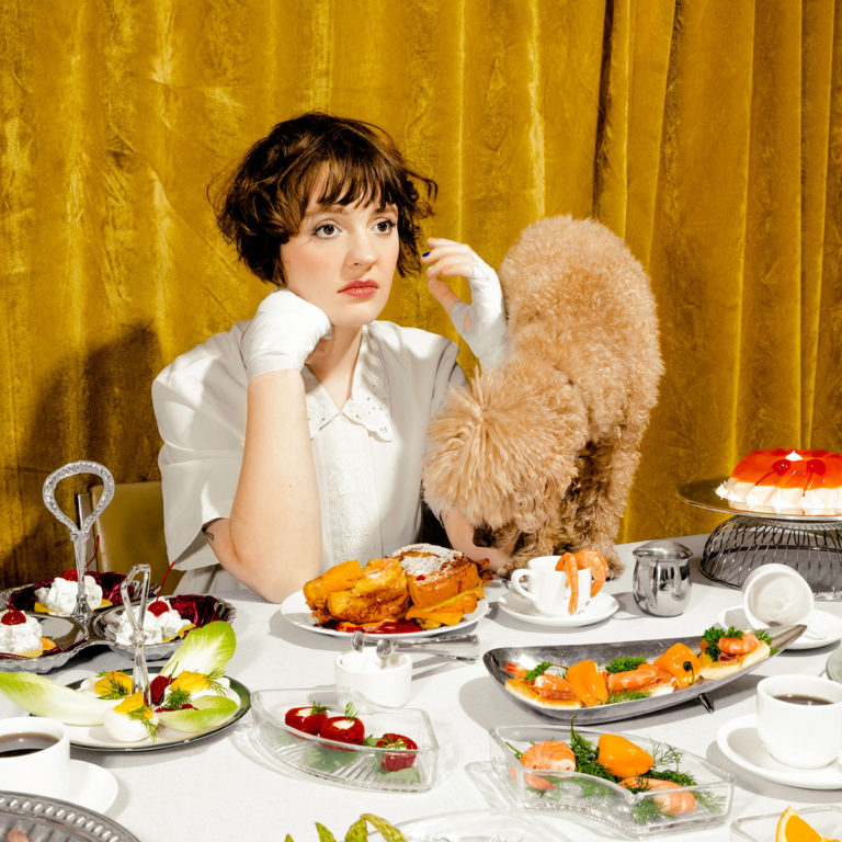 Eine junge Frau sitzt gelangweilt an einem opulent gedeckten Tisch mit Retro-Essen, während ein Pudel ihren Teller ableckt. Vor goldgelbem Vorhang, inszeniert wie ein absurdes Stillleben.
