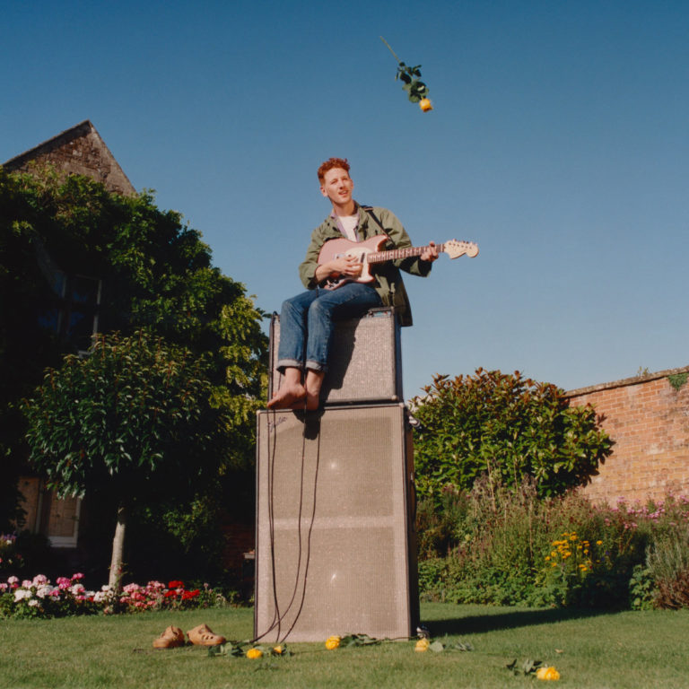 Person sitzt mit Gitarre auf Verstärkern im Garten, umgeben von Blumen.