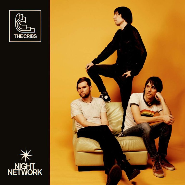 Drei Männer von The Cribs posieren vor orangefarbenem Hintergrund auf einem Sofa.