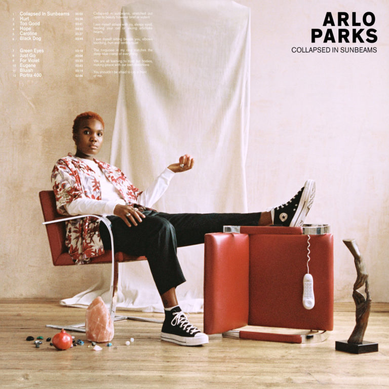 Albumcover von Arlo Parks „Collapsed in Sunbeams“ zeigt die Sängerin lässig auf einem roten Stuhl sitzend, umgeben von Alltagsobjekten in minimalistischer Kulisse.