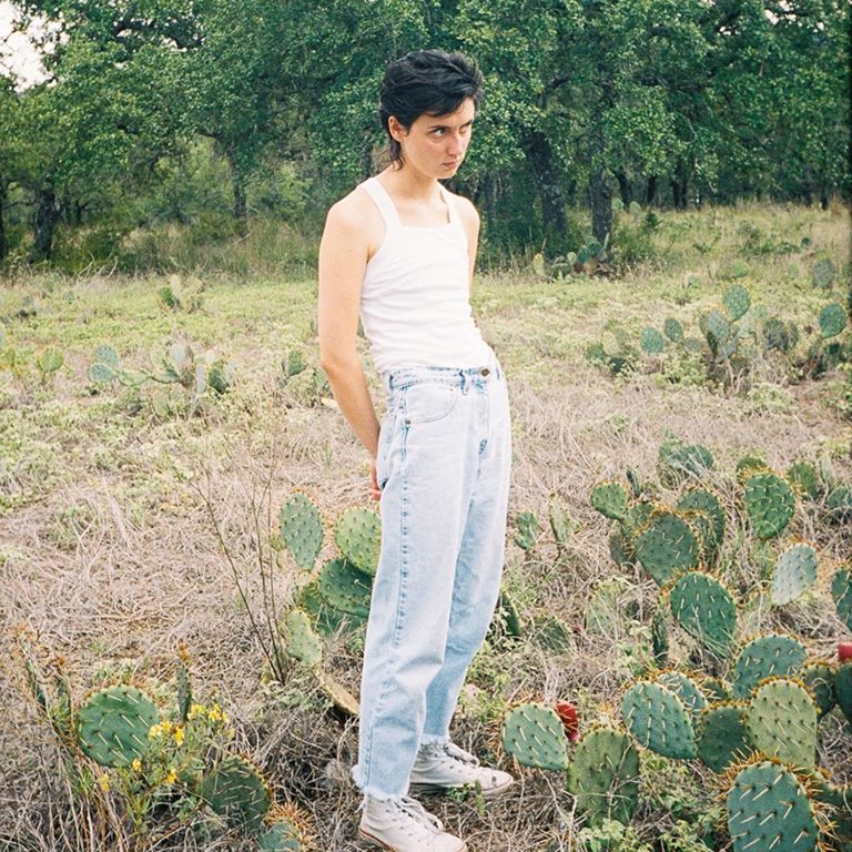 Eine junge Frau mit weißem Tanktop und hellblauer Jeans steht zwischen Kakteen in einer texanischen Wildnis und blickt herausfordernd in die Kamera.