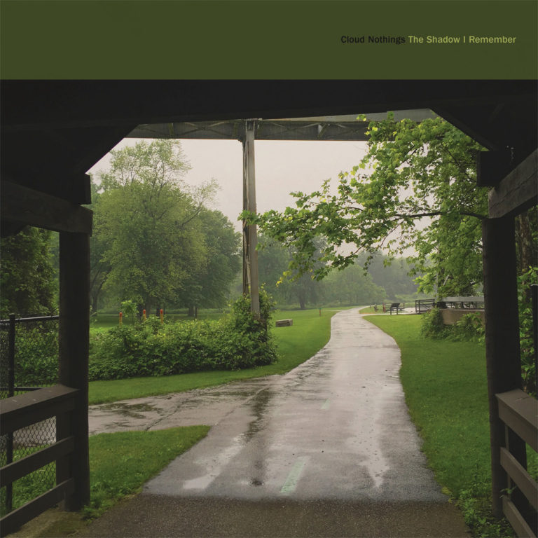 Ein verregneter Parkweg, aufgenommen unter einem Holzunterstand, führt durch eine grüne Landschaft im Nebel. Das Setting wirkt ruhig, introspektiv und leicht melancholisch.