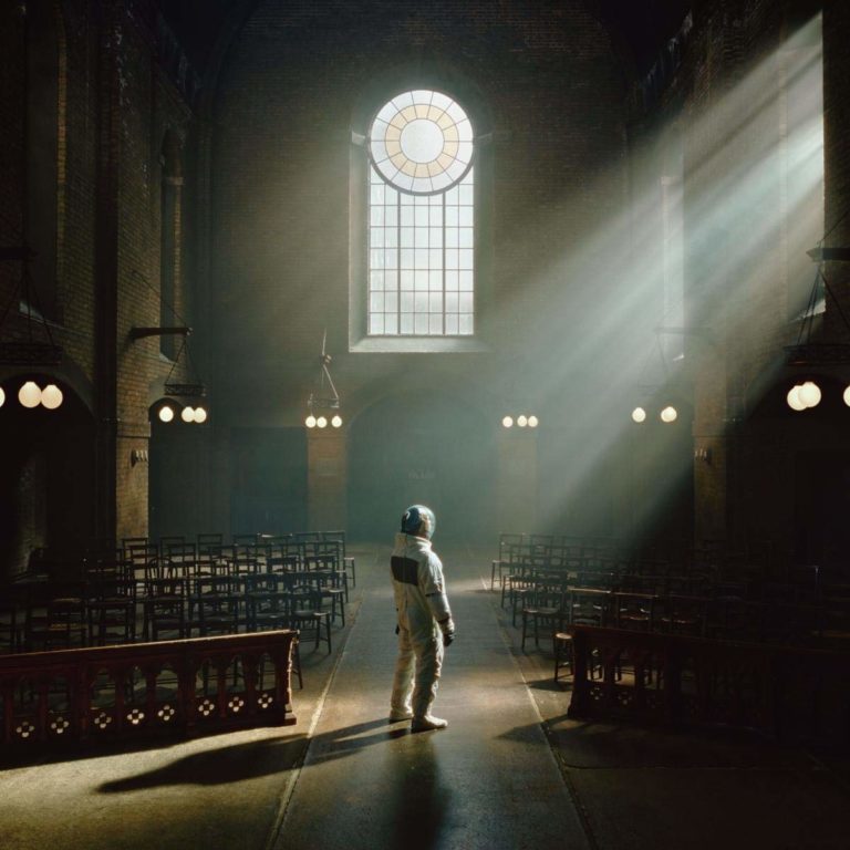 Ein einzelner Astronaut steht in einer leeren, lichtdurchfluteten Kirche. Sonnenstrahlen brechen durch ein großes Rundfenster – surreal, kontemplativ, erhaben.