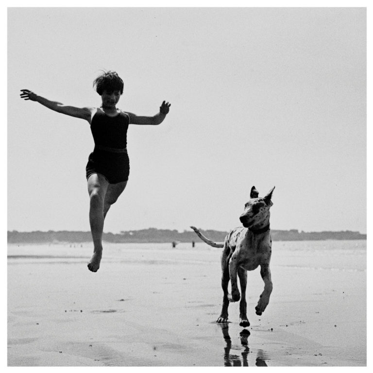 Ein Mädchen in Badekleidung springt übermütig am Strand, neben ihr rennt ein großer gefleckter Hund – eingefangen in schwarz-weiß nostalgischer Leichtigkeit.