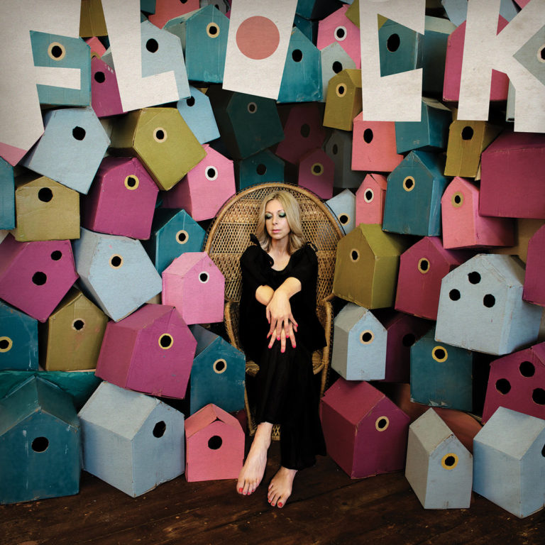 Jane Weaver sitzt barfuß in einem Korbstuhl vor einer Wand aus bunten Vogelhäuschen in Blau, Pink, Grün und Violett. Über ihr hängen große Buchstaben, die das Wort „FLOCK“ bilden. Das Setting ist surreal, verspielt und grafisch inszeniert – ein Kunst-Statement zwischen Retro-Charme und eigenwilliger Pop-Ästhetik.