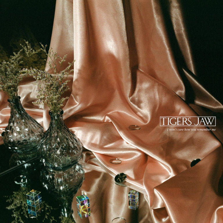 Albumcover von I Won’t Care How You Remember Me der Band Tigers Jaw: Eine Szene mit roséfarbenem, glänzendem Stoff, auf dem kleine Glasringe, Spiegel und zwei Kristallvasen mit Trockenblumen arrangiert sind. Das Bild wirkt nostalgisch, zart und zugleich leicht surreal durch die Spiegelungen und Lichtreflexe. Der Albumtitel ist dezent in einem weißen Rahmen oben rechts eingeblendet.