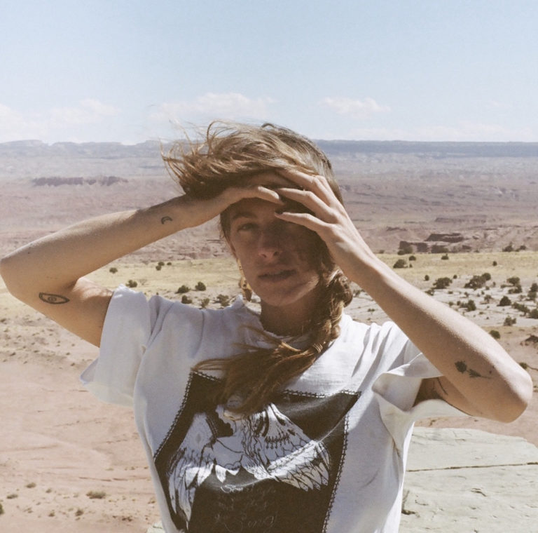 Indigo Sparke steht auf einem Felsplateau in weiter, karger Wüstenlandschaft. Der Wind weht ihr ins Gesicht, während sie eine Hand schützend über die Augen hält. Sie trägt ein weißes T-Shirt mit grafischem Druck. Die Szenerie wirkt introspektiv, rau und weit – eine stimmungsvolle Verbindung aus Natur, Verletzlichkeit und innerer Stärke.