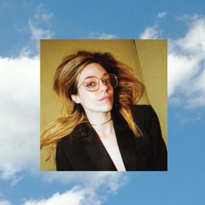 Cover von Anna Fox Rochinskis Album Cherry: Die Künstlerin liegt in Blazer und Brille auf hellem Untergrund, das Bild schwebt vor blauem Himmel mit Wolken.
