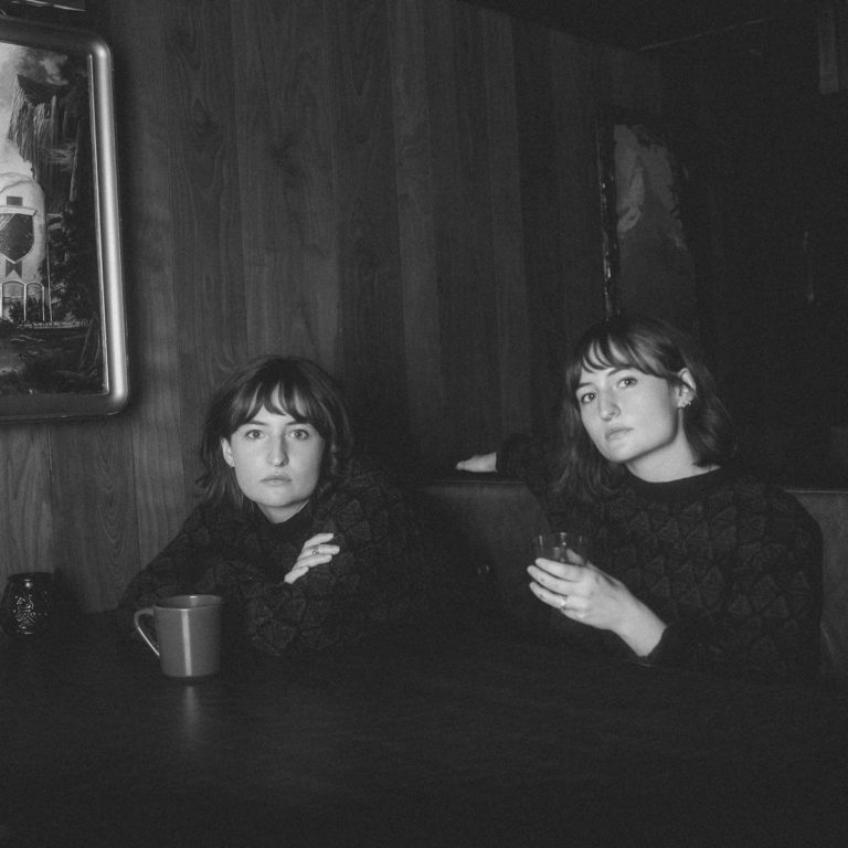 Schwarz-weißes Foto von zwei Frauen in identischer Kleidung an einem dunklen Holztisch, eine mit Kaffeetasse, die andere mit Glas – melancholische Barstimmung