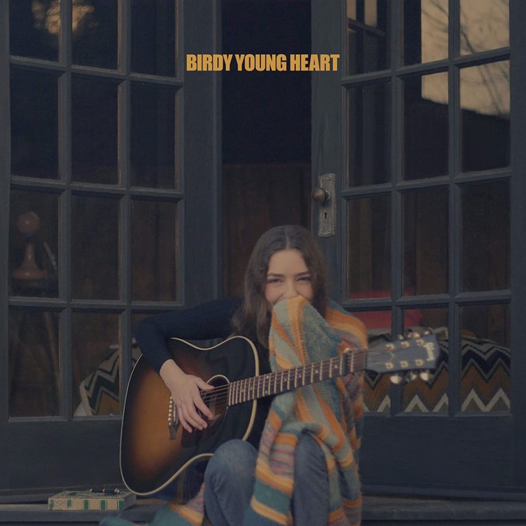 Birdy sitzt auf einer Veranda vor einer dunklen Holztür, umhüllt von einer bunten Decke, mit Akustikgitarre im Schoß und einem scheuen Lächeln im Gesicht. Der Schriftzug „Birdy Young Heart“ erscheint oben in gelber Schrift.