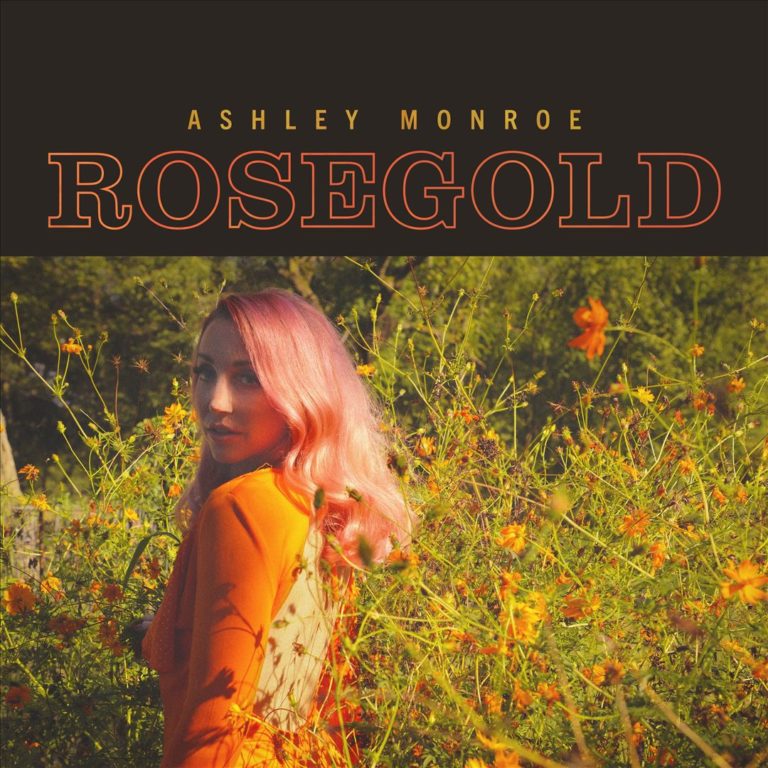 Ashley Monroe steht mit roséfarbenen Haaren und orangefarbenem Kleid inmitten eines Feldes voller gelber Wildblumen. Der obere Teil des Covers zeigt in orange-gelber Typografie den Titel „Rosegold“.
