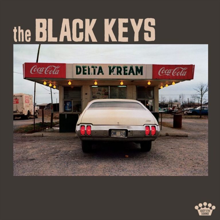 Ein weißer Oldtimer steht vor einem klassischen amerikanischen Diner mit dem Schild "Delta Kream", eingerahmt von Coca-Cola-Schriftzügen. Das Bild wirkt retro und rustikal.