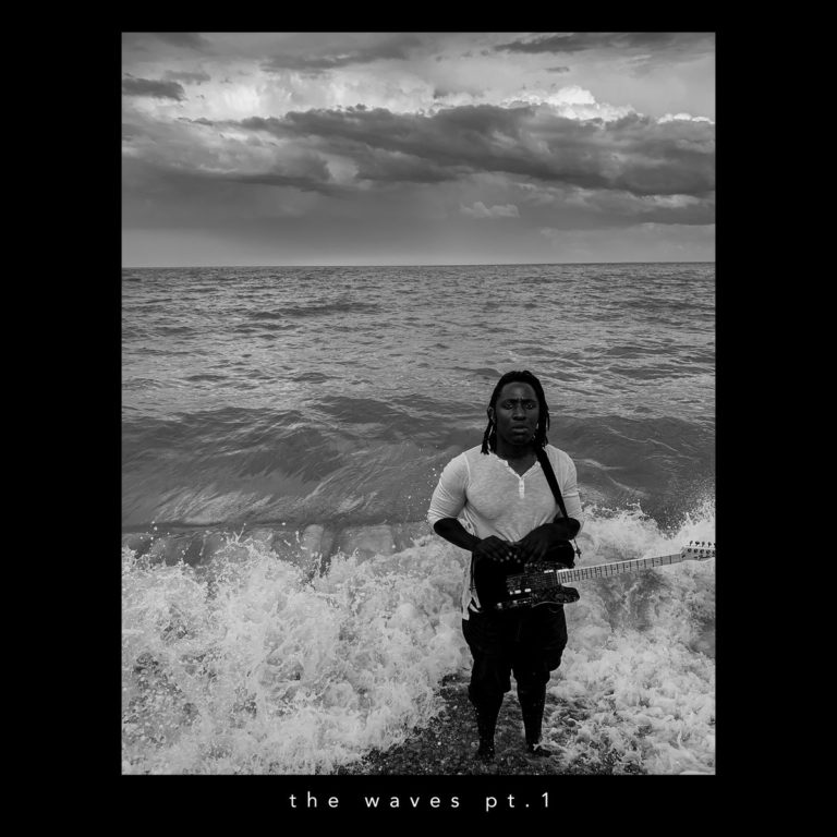 Schwarzweißbild von Kele Okereke mit Gitarre am Meer, umgeben von aufspritzenden Wellen und dramatischem Himmel – gerahmt von schwarzem Rand.