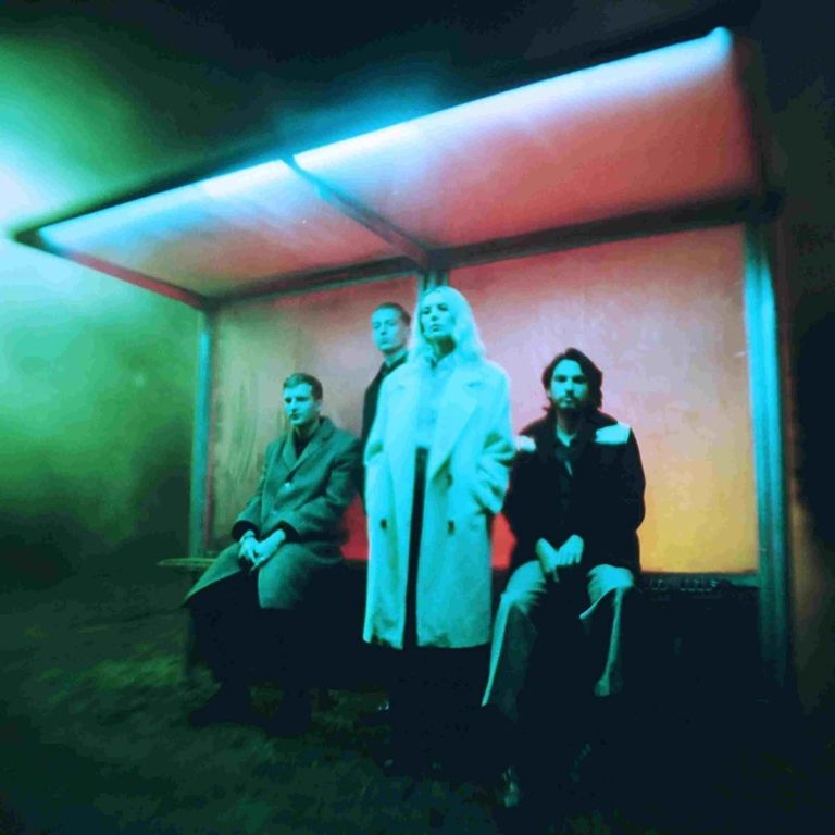 Die Band Wolf Alice sitzt in einer leuchtenden Bushaltestelle bei Nacht, umgeben von Nebel und kaltem Licht in Cyan, Grün und Magenta.