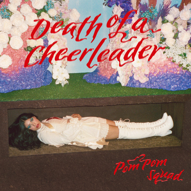 Das Albumcover von „Death of a Cheerleader“ von Pom Pom Squad zeigt eine Frau im Cheerleader-Outfit, die in einem offenen Sarg unter einer künstlich-blumigen Kulisse liegt – zwischen Camp, Kritik und Glamour inszeniert.