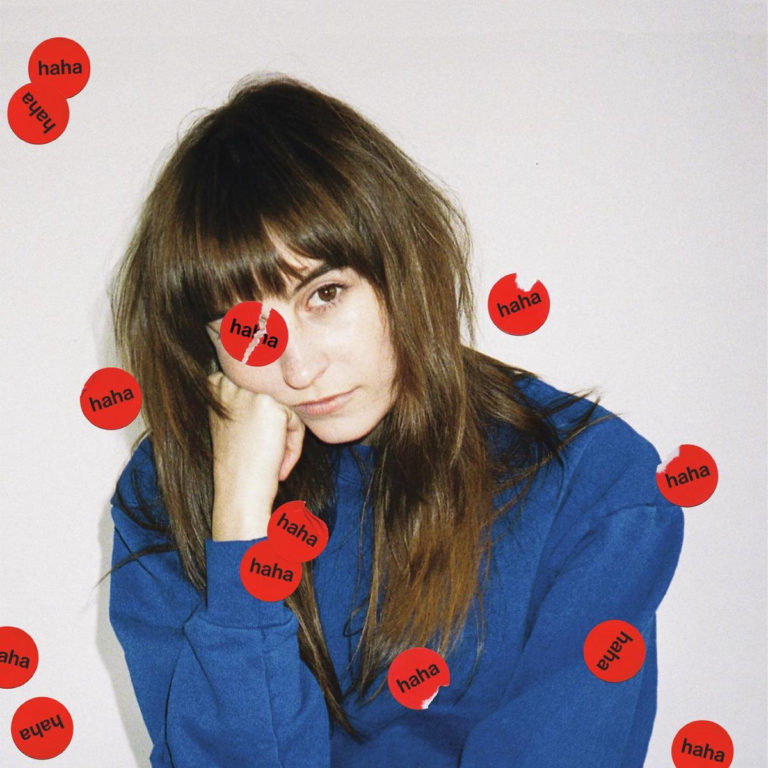 Das Albumcover von „I Know I’m Funny haha“ von Faye Webster zeigt sie mit gelangweiltem Blick in einem blauen Sweater, umgeben von roten Aufklebern mit der Aufschrift „haha“.