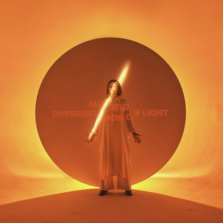 Jade Bird steht in einem leuchtenden orangen Kreis, durchdrungen von einem Lichtstrahl – wie eine Heldin in einer Retro-Zeitkapsel aus Gefühl und Klang.