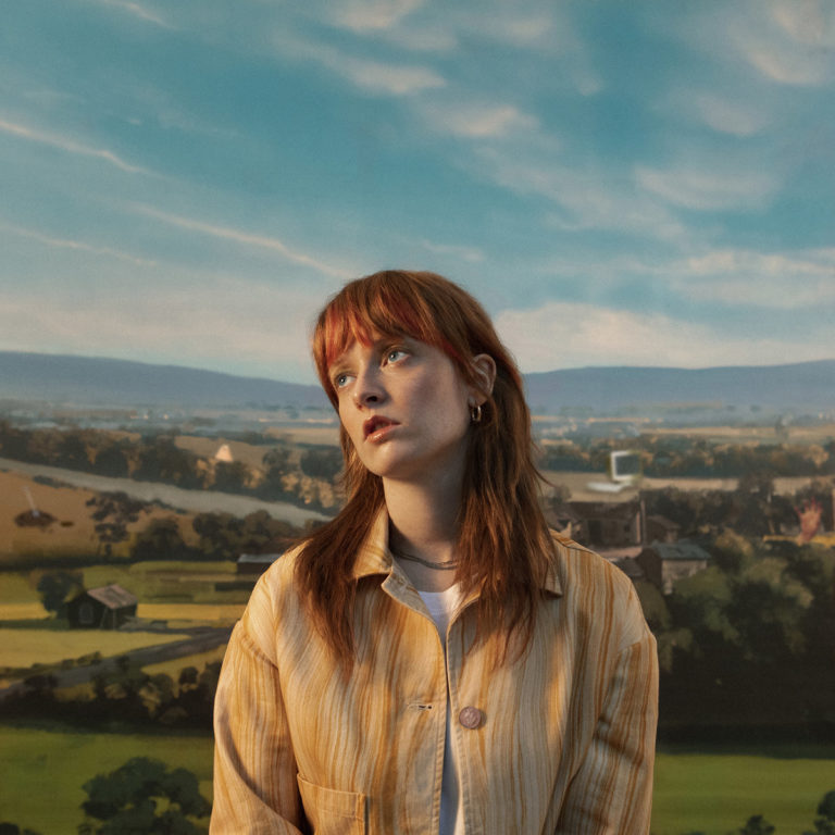 Orla Gartland steht gedankenverloren vor einem gemalten Landschaftspanorama – zwischen Idylle, Melancholie und innerem Aufruhr.