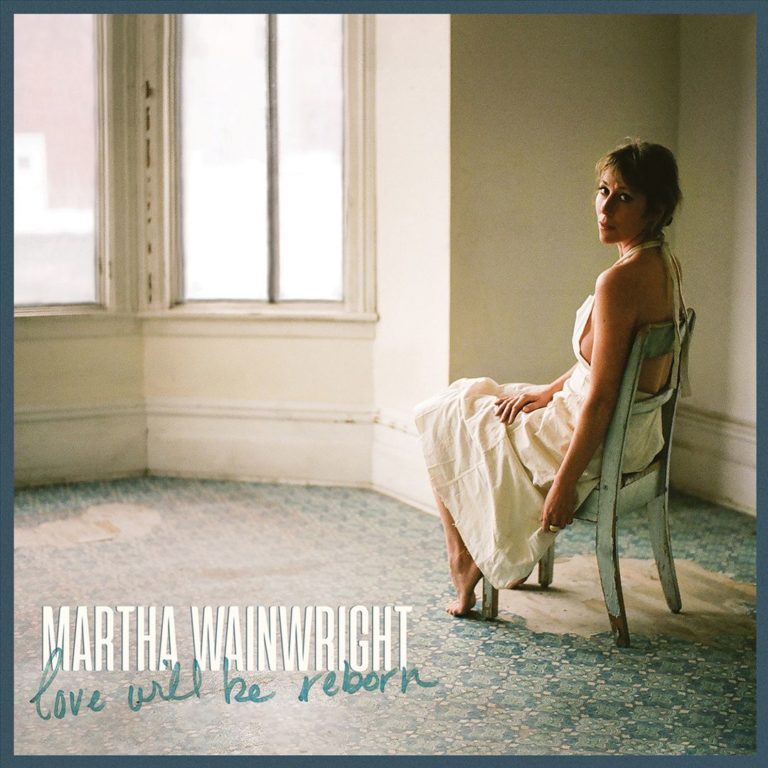 Martha Wainwright sitzt barfuß und in einem cremefarbenen Kleid auf einem abgenutzten Stuhl in einem leeren, lichtdurchfluteten Raum. Der Boden ist alt, das Fenster groß, der Blick ruhig und direkt – eine Szene voller melancholischer Würde.