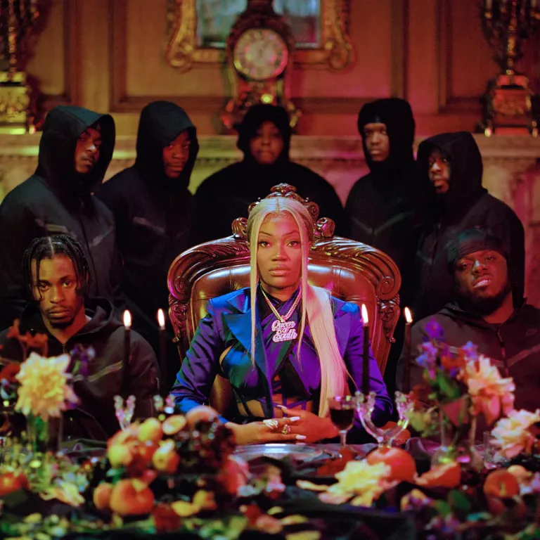 Albumcover zu Queen of the South: Shaybo sitzt in königlicher Pose an einer prunkvoll gedeckten Tafel, flankiert von Männern in schwarzen Hoodies, im luxuriös ausgeleuchteten Raum.