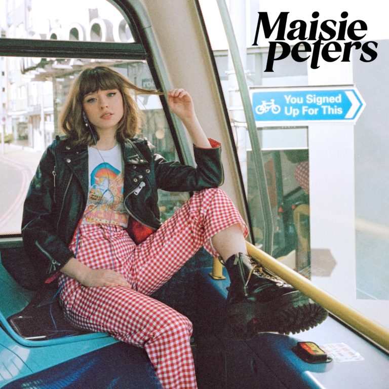 Maisie Peters sitzt lässig in einem Doppeldeckerbus, trägt karierte Hose, Bandshirt und Lederjacke. Neben ihr ein Straßenschild: „You Signed Up For This“. Das Bild wirkt retro, urban und charmant inszeniert – ganz im Stil eines Coming-of-Age-Albums.