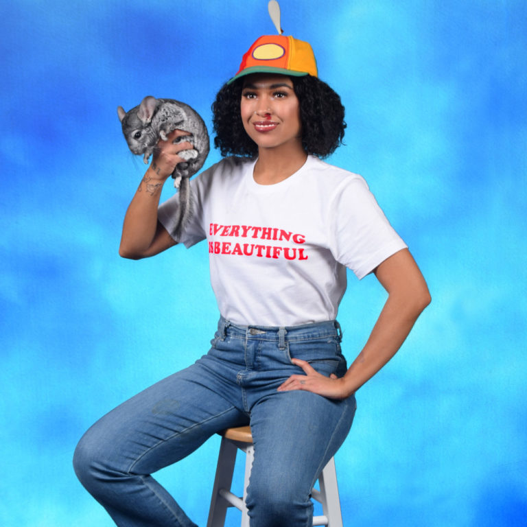 Princess Nokia posiert lässig auf einem Hocker vor blauem Hintergrund, trägt ein weißes Shirt mit rotem Schriftzug „Everything Is Beautiful“, eine bunte Mütze und hält ein kleines graues Chinchilla.