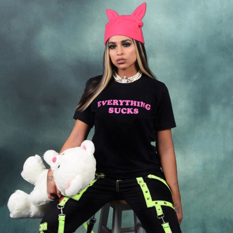Princess Nokia sitzt vor grauem Hintergrund auf einem schwarzen Hocker, trägt pinke Mütze mit Katzenohren, schwarzes T-Shirt „Everything Sucks“ und neongelbe Details, hält ein weißes Stofftier.