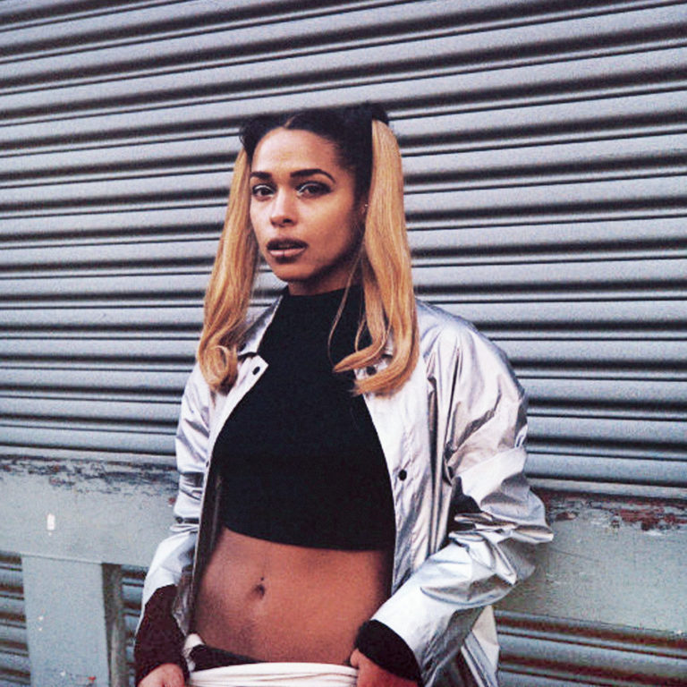 Princess Nokia steht vor einer grauen Metall-Rolltorwand, trägt silberne Jacke, schwarzen Crop-Top und blonde Zöpfe, wirkt selbstbewusst und urban.