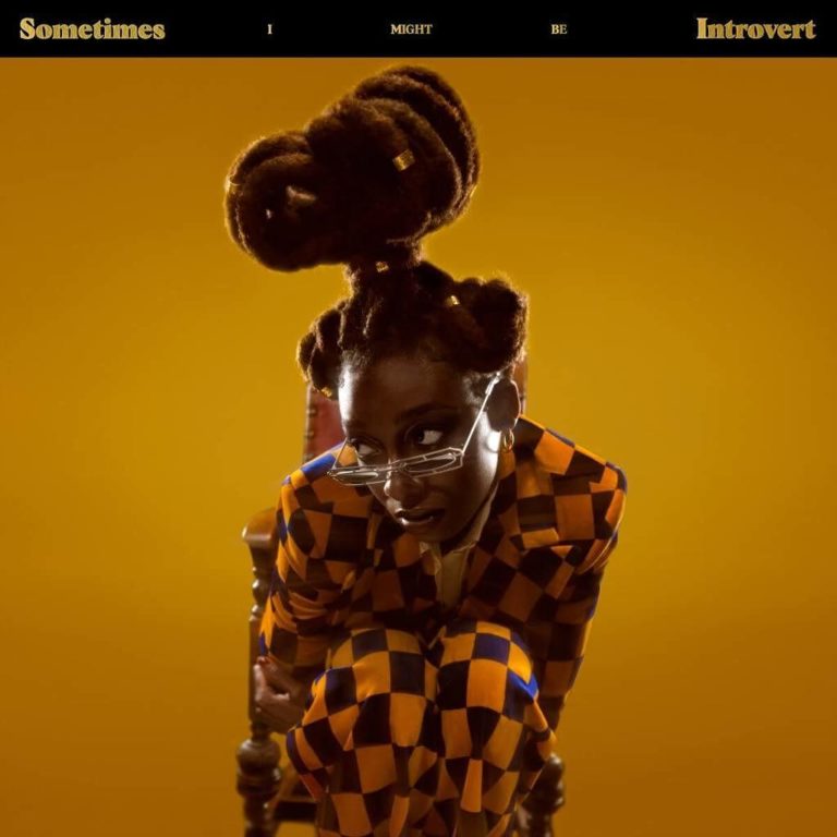 Coverbild von „Sometimes I Might Be Introvert“: Little Simz hockt auf einem Stuhl vor gelbem Hintergrund, trägt einen orange-blauen Karoanzug und schaut über eine schmale Brille direkt in die Kamera. Ihre Frisur ist kunstvoll hochgesteckt.