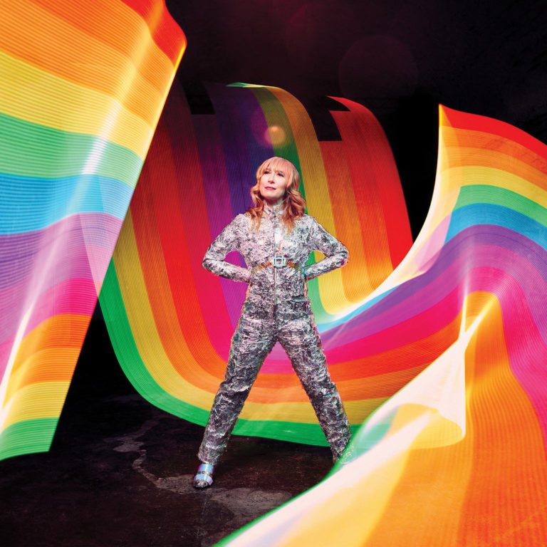 Erika Wennerstrom im silbernen Glitzeranzug vor wirbelnden Regenbogenbändern auf schwarzem Hintergrund – ein lebendiges, hoffnungsvolles Artwork voller Energie.