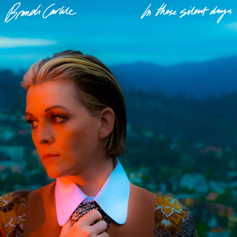 Cover von „In These Silent Days“ von Brandi Carlile: Die Künstlerin blickt ernst nach links, in warmem Sonnenlicht vor kühlem blauen Abendhimmel und Hügellandschaft.