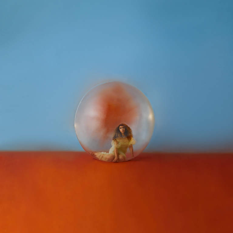 Cover von „In The Meantime“: Alessia Cara sitzt klein in einer gläsernen Kugel auf oranger Fläche vor himmelblauem Hintergrund – minimalistisch und surreal.