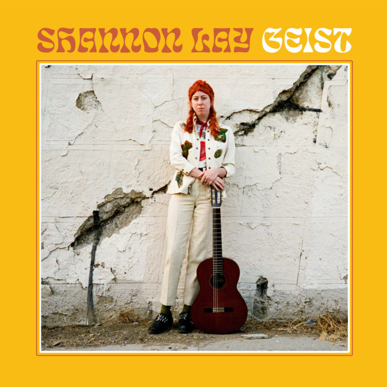 Albumcover von Shannon Lay – „Geist“: Die Musikerin lehnt mit roter Mütze und Akustikgitarre an einer bröckelnden Wand, eingerahmt von einem gelben Rand.