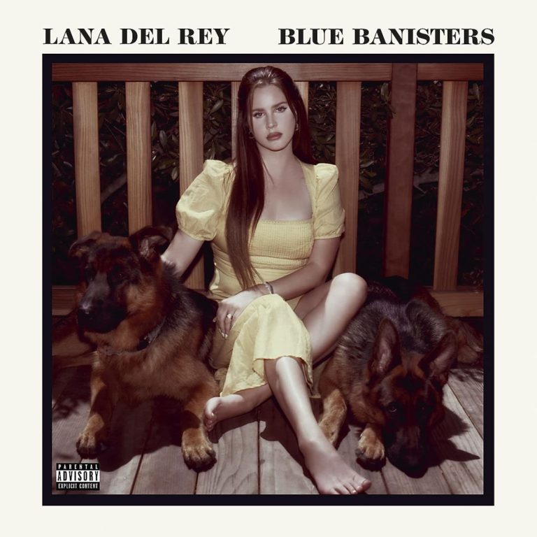 Lana Del Rey sitzt barfuß in einem gelben Kleid auf einer Holzveranda zwischen zwei Schäferhunden – ein stilles, melancholisches Porträt.
