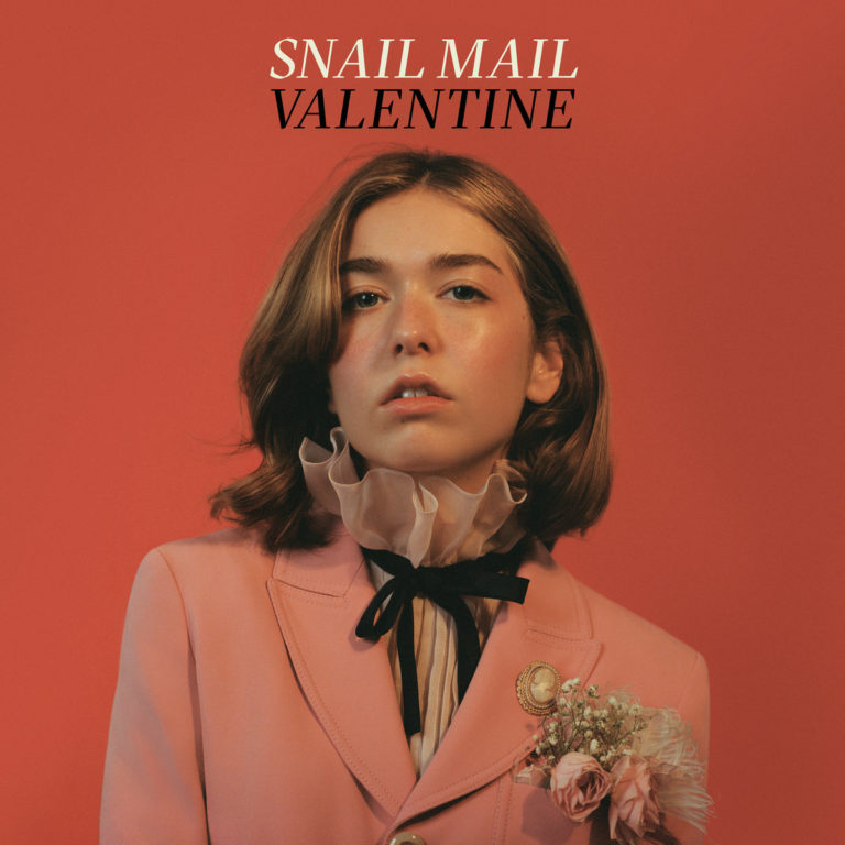 Lindsey Jordan alias Snail Mail trägt einen rosafarbenen Blazer mit Brosche und Rüschenkragen vor rotem Hintergrund – Cover des Albums „Valentine“.