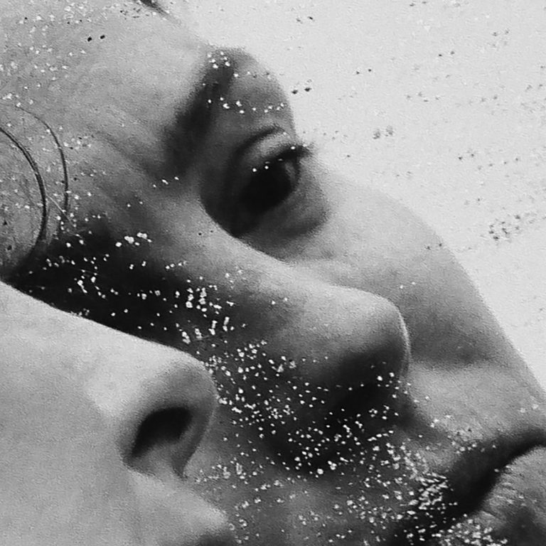 Ein schwarz-weißes Close-up zweier Gesichter, eins davon halb verdeckt und von Glitzerstaub überzogen – verletzlich, fragmentiert, fast geisterhaft.